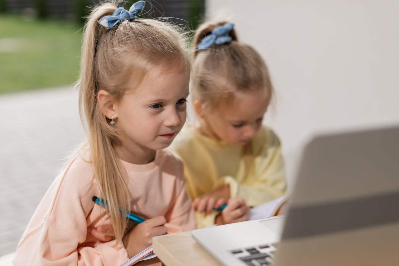 Two young girls sitting outdoors focused on online learning using a laptop, writing and participating in a virtual class.